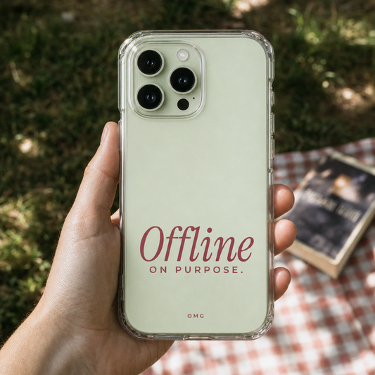 Clear iPhone case printed “Offline on purpose.” in dark rose, held over a picnic blanket
