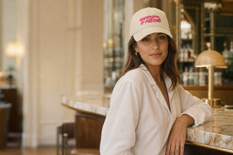 Ivory classic cap embroidered “expensive & difficult” in pink, worn at a marble hotel bar