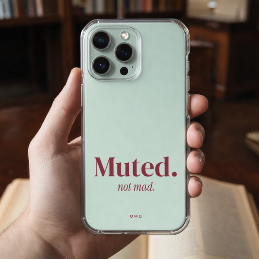 Clear iPhone case printed “Muted. not mad.” in dark rose, held in one hand at a library desk